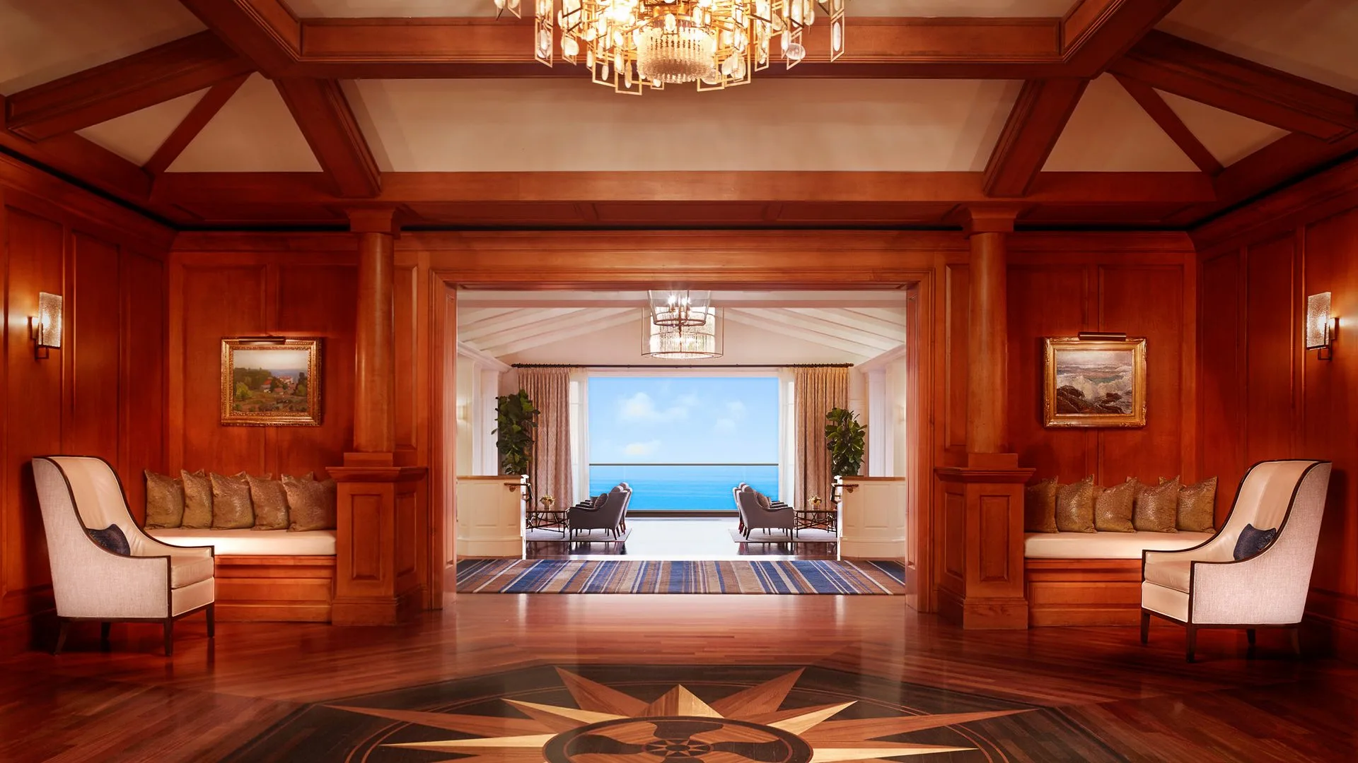 Elegant wood-paneled lounge opening to ocean view, with chandeliers, seating alcoves, and symmetrical design.