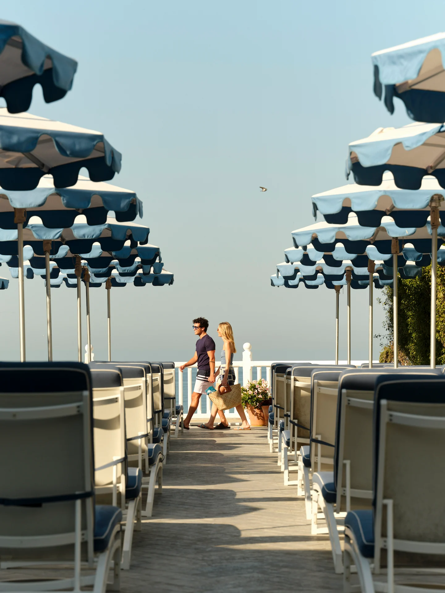 Beachside walkway lined with lounge chairs and striped umbrellas, couple walking toward ocean view under sunny sky.