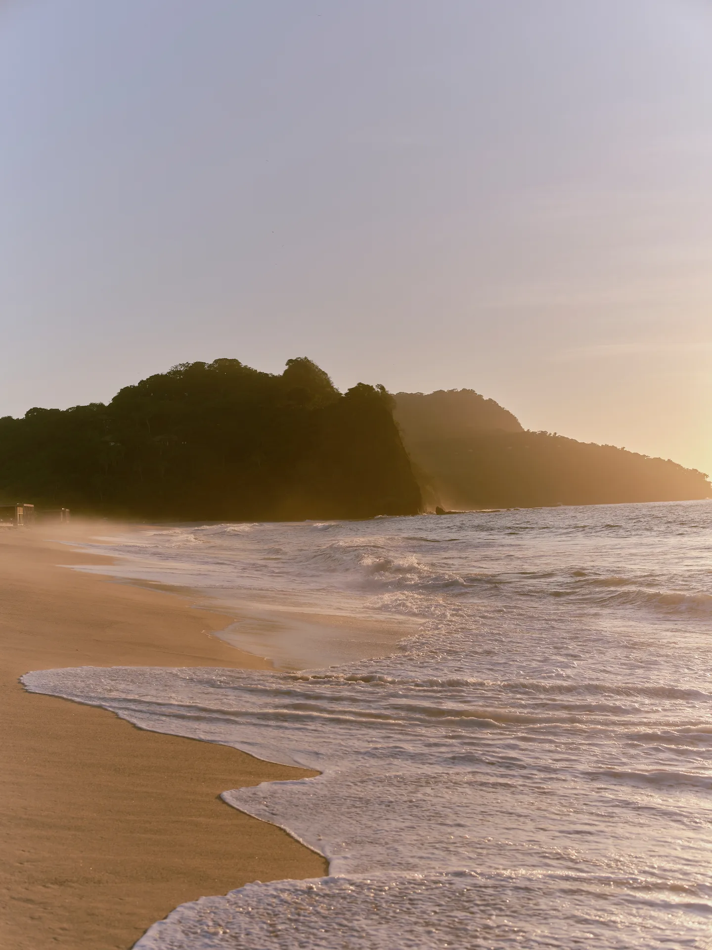 Golden hour waves washing onto a quiet sandy beach with forested cliffs and soft sunlight in the distance.