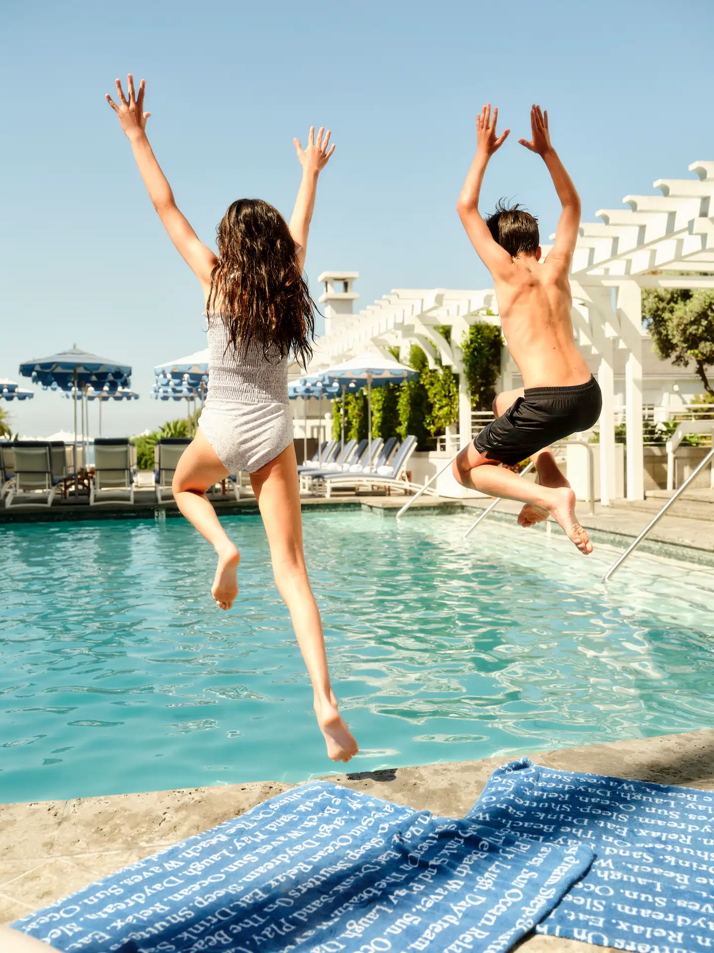Two people mid-jump into sunlit pool, surrounded by striped umbrellas, lounge chairs, and playful summer vibes.