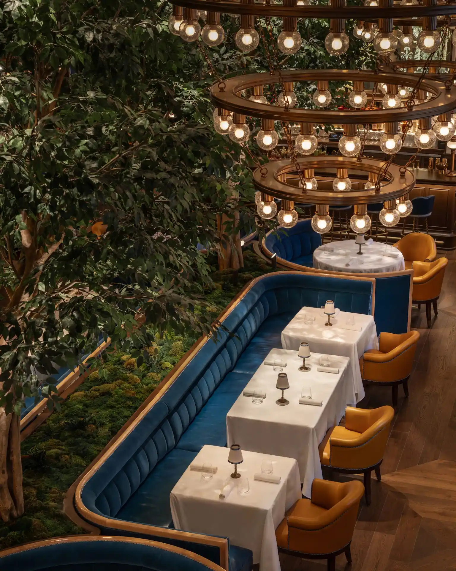 Upscale restaurant with blue banquettes, yellow armchairs, tiered chandelier, and lush greenery.