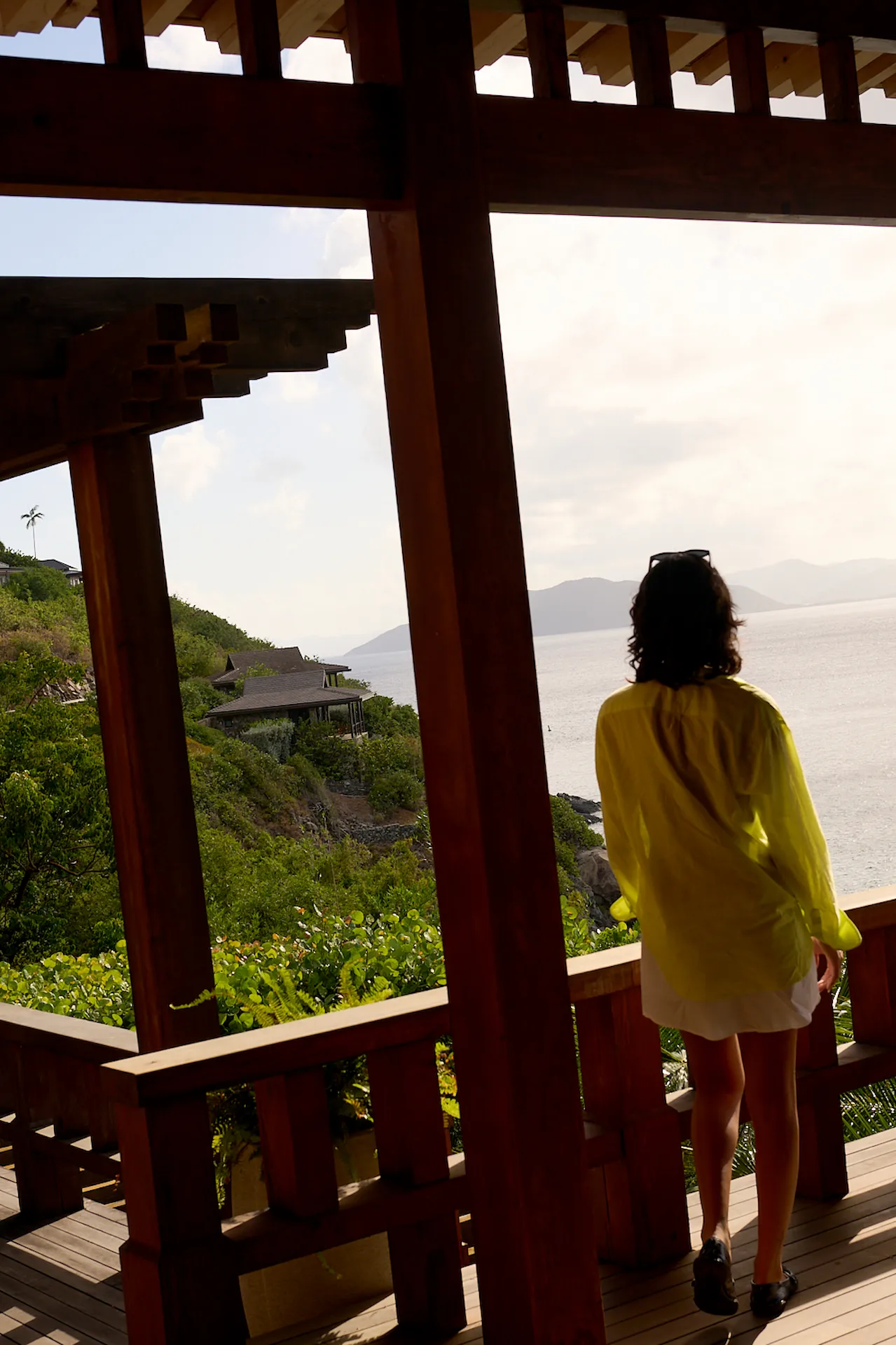 Person in yellow cover-up on a wooden balcony overlooking the ocean, lush green hills, and coastal villas.