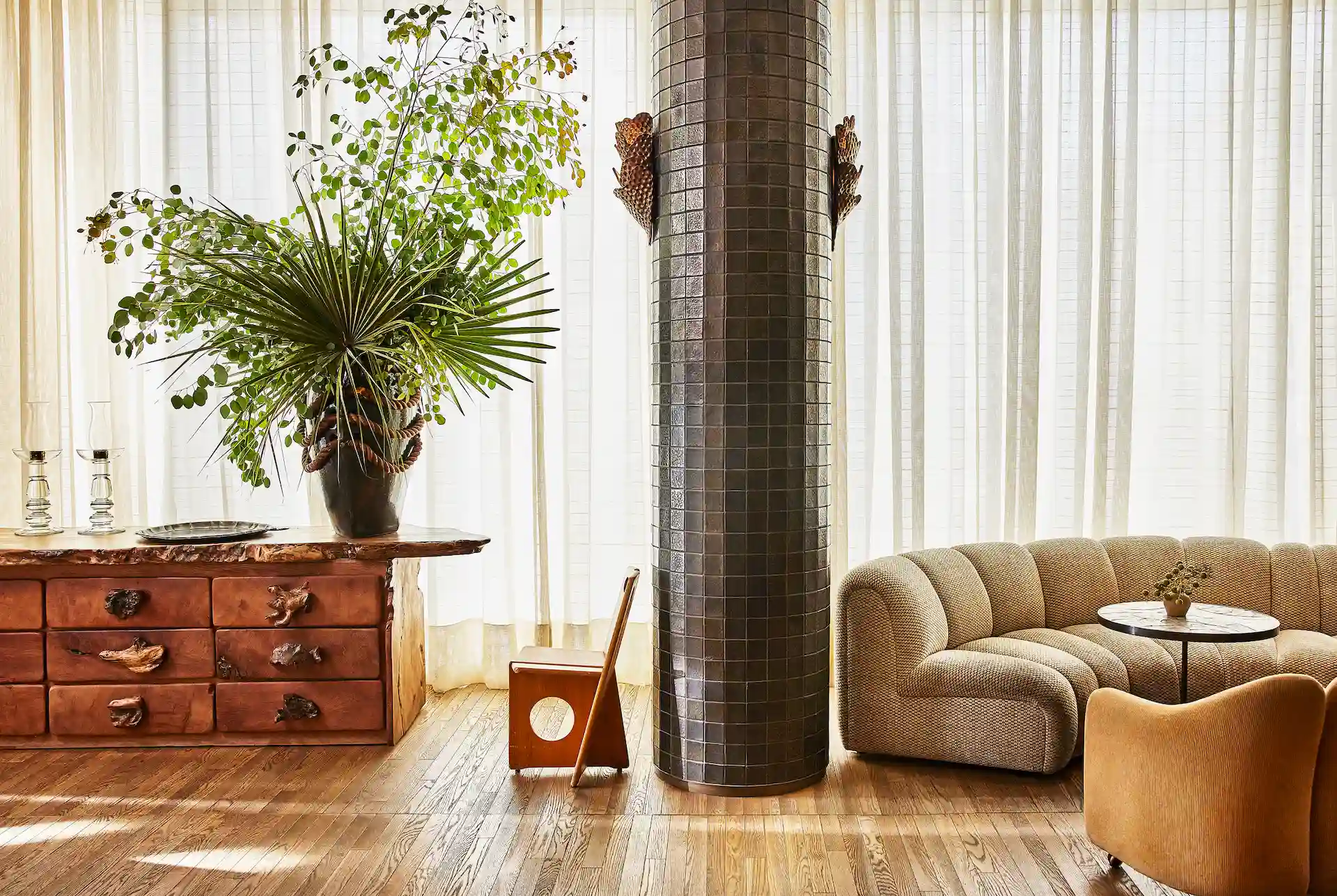 Hamak 3 Sunlit lounge with curved sofa, tiled column, wooden floors, vintage cabinet, and lush indoor plants by sheer curtains.