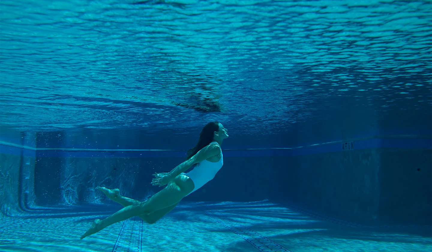 Woman swimming underwater in pool, sunlight rippling across blue water surface.