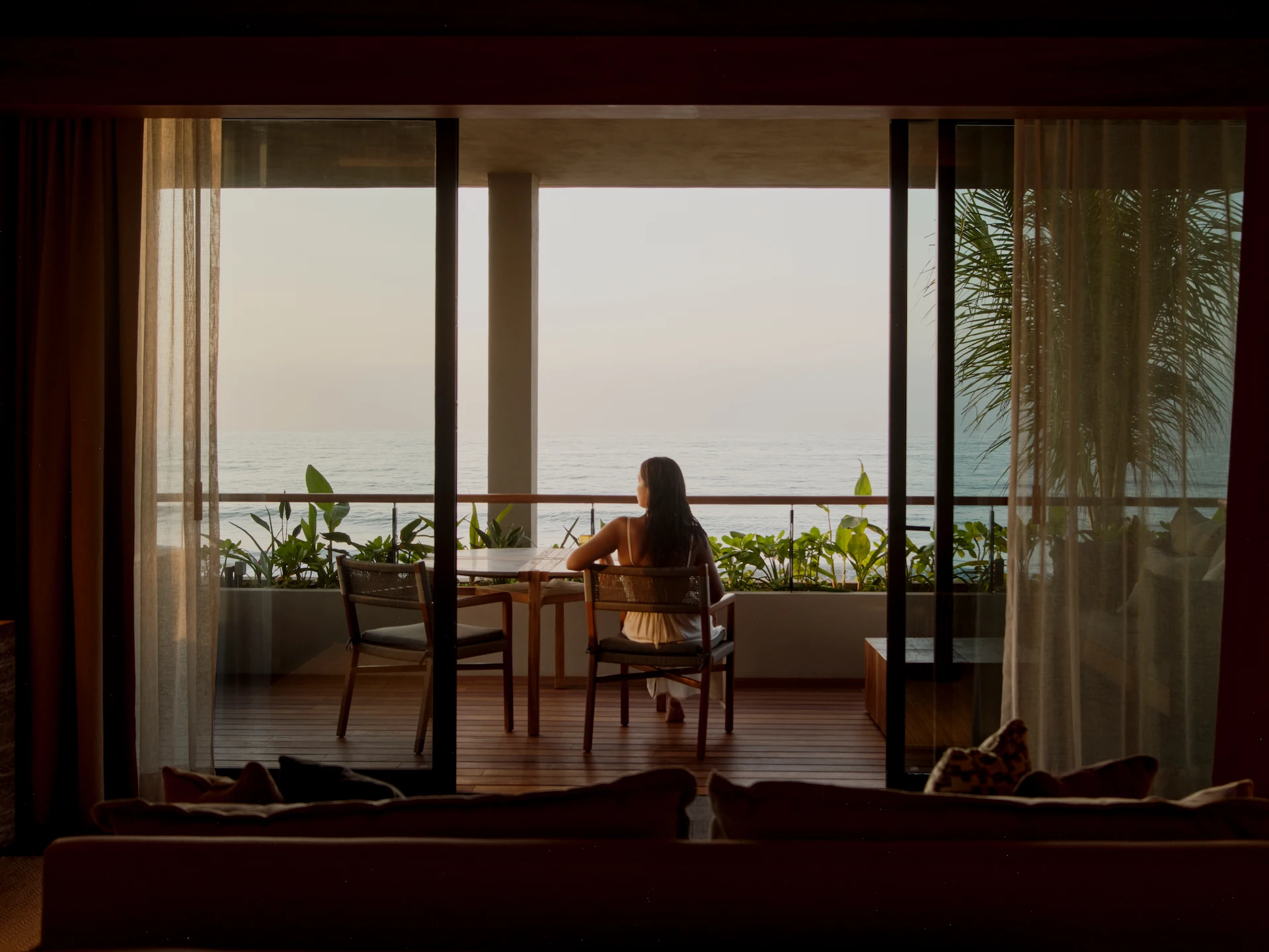 Oceanview balcony at Rosewood Kona Village with guest seated at wooden table, framed by lush greenery and sheer curtains.