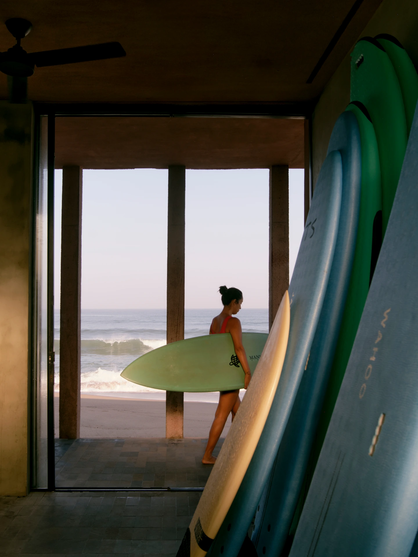 Guest walking barefoot with surfboard toward ocean at Rosewood Mandarina, framed by wooden beams and stacked boards.