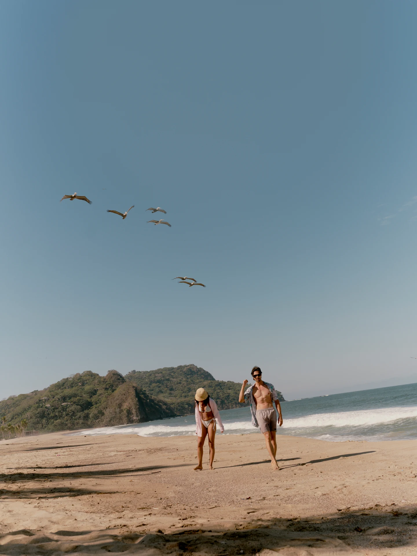Barefoot guests walking along the beach at Rosewood Mandarina, birds soaring overhead and lush hillside beyond.