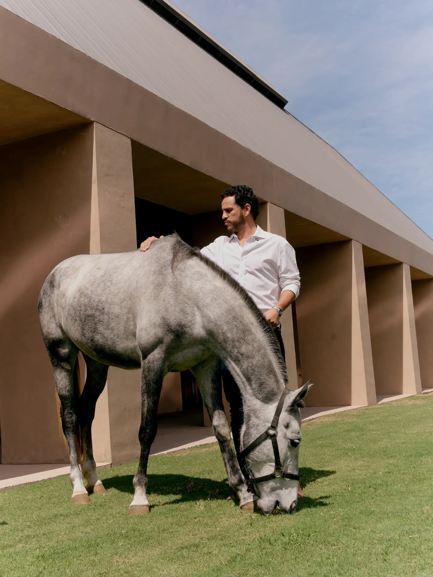 Man standing beside gray horse on lawn at Rosewood Mandarina, modern architecture and clear sky in background.
