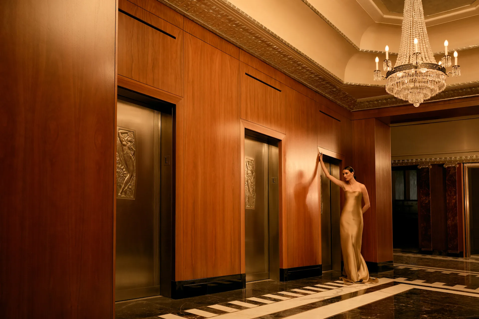 Elegant elevator lobby at Waldorf Astoria New York with sculpted bronze panels, marble floors, and guest in gold dress.