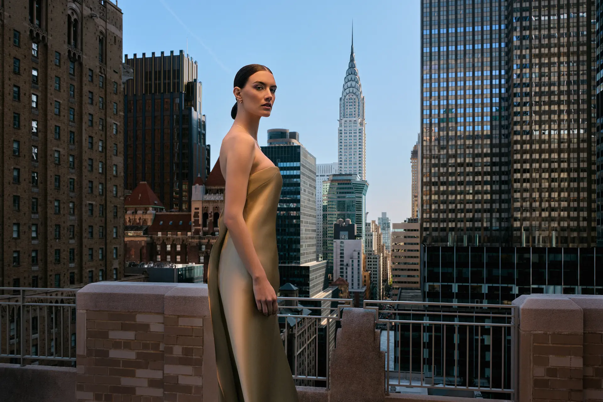 Person in gold strapless gown standing on rooftop terrace with city skyline and Chrysler Building behind.