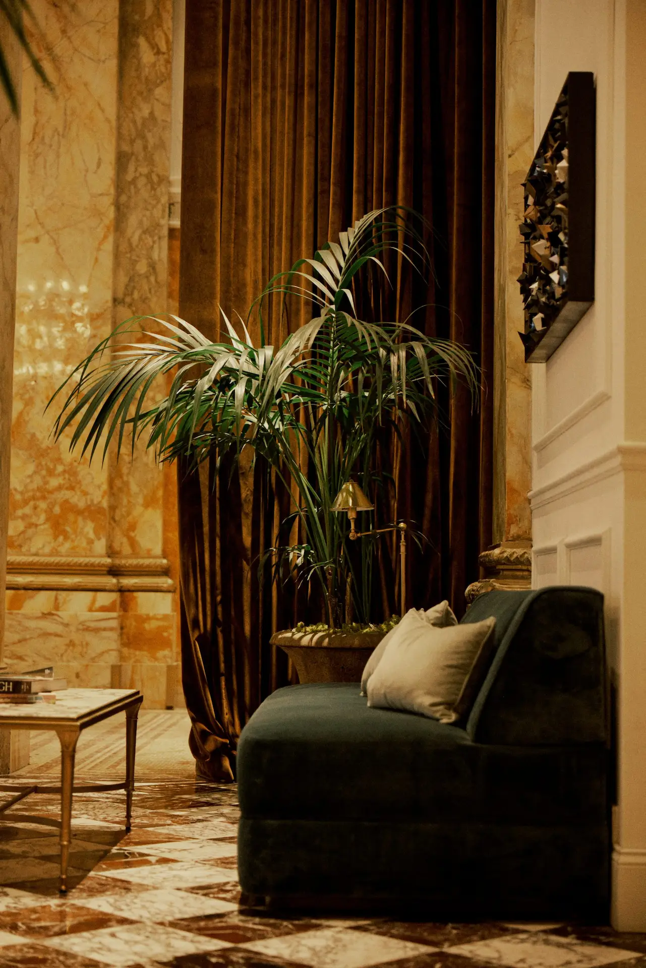 Velvet chaise lounge with throw pillows, marble floor, potted palm, and ornate wall decor.