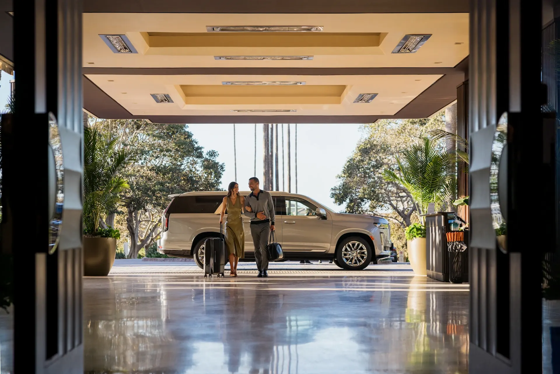 Couple arriving at a luxury hotel entrance with luggage, parked SUV behind, and sunlit driveway under canopy.