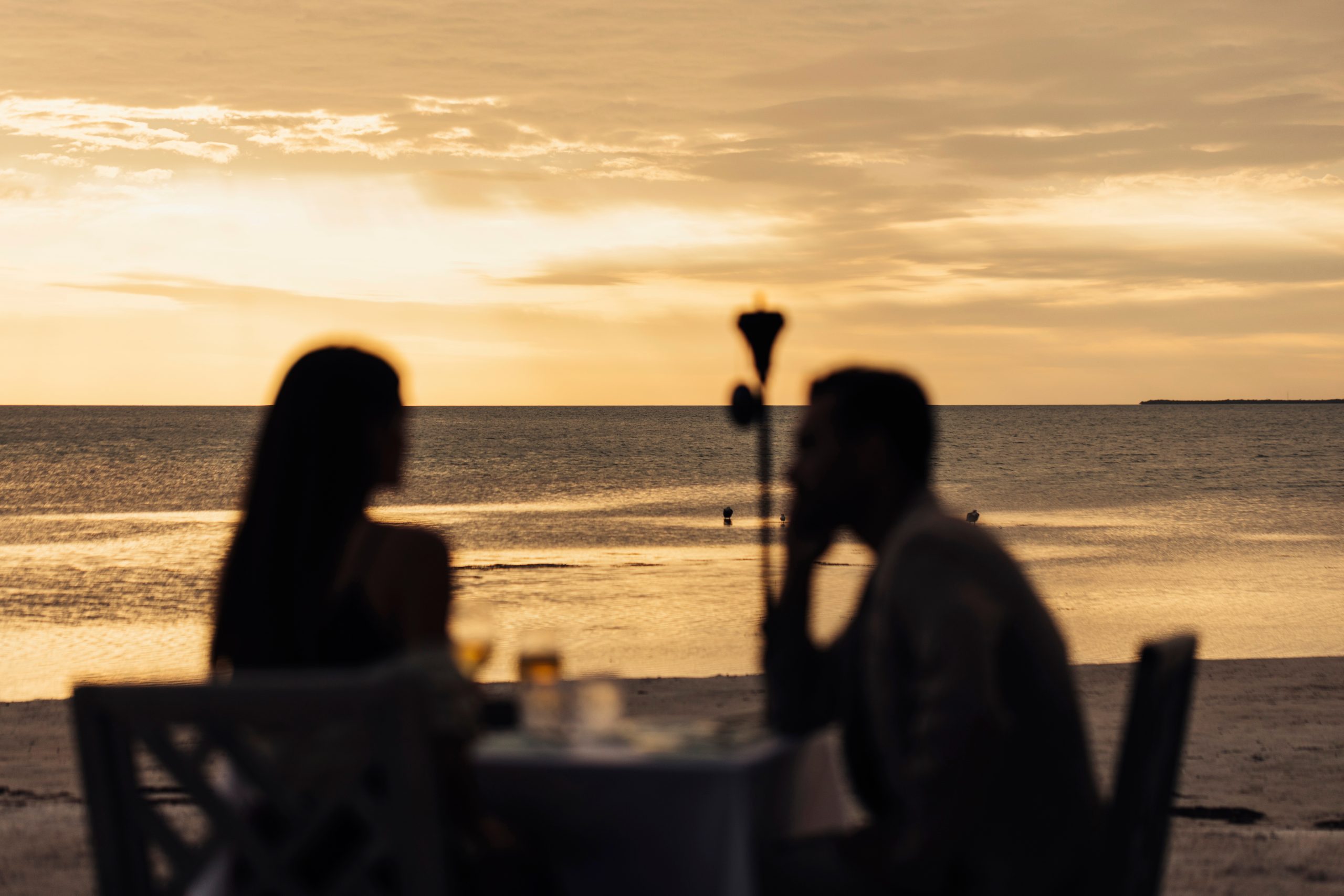 Silhouetted couple dining at a beach table during golden sunset, with calm ocean and clouds in the background.