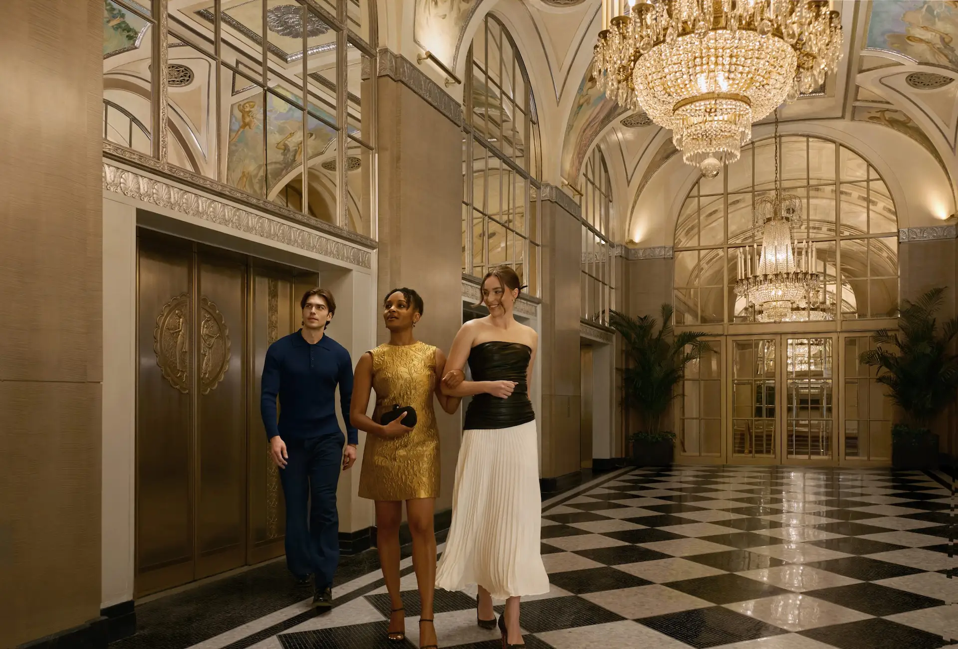 Three people walk through ornate hotel lobby with chandeliers, marble floor, and decorative ceiling.
