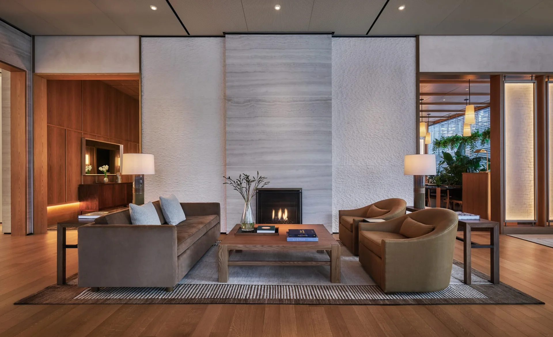 Modern hotel lobby lounge with fireplace, neutral sofas, wooden accents, soft lighting, and indoor plants.