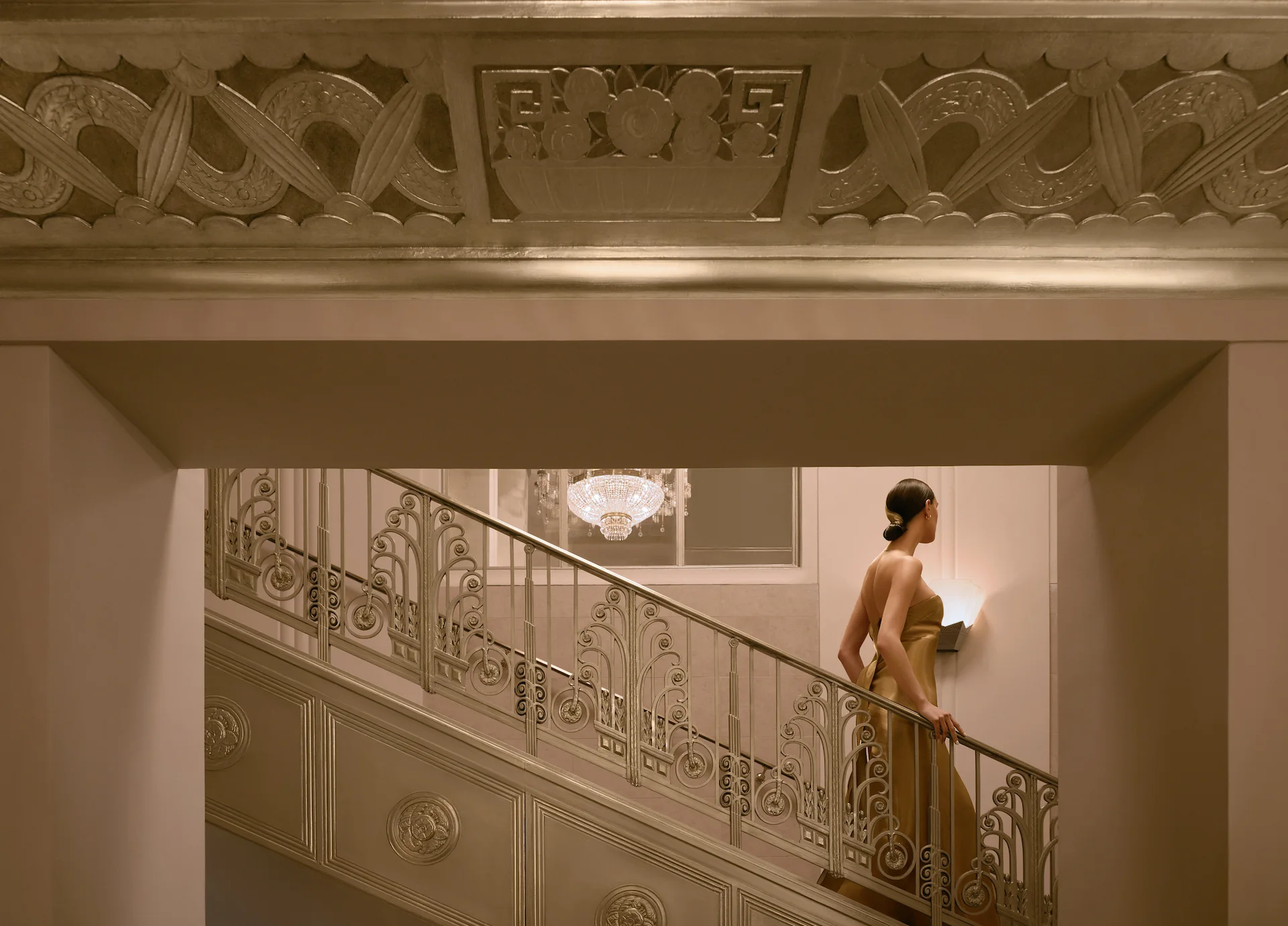 Woman in an elegant gown ascending a grand staircase with ornate railings, chandelier, and classical interior details.