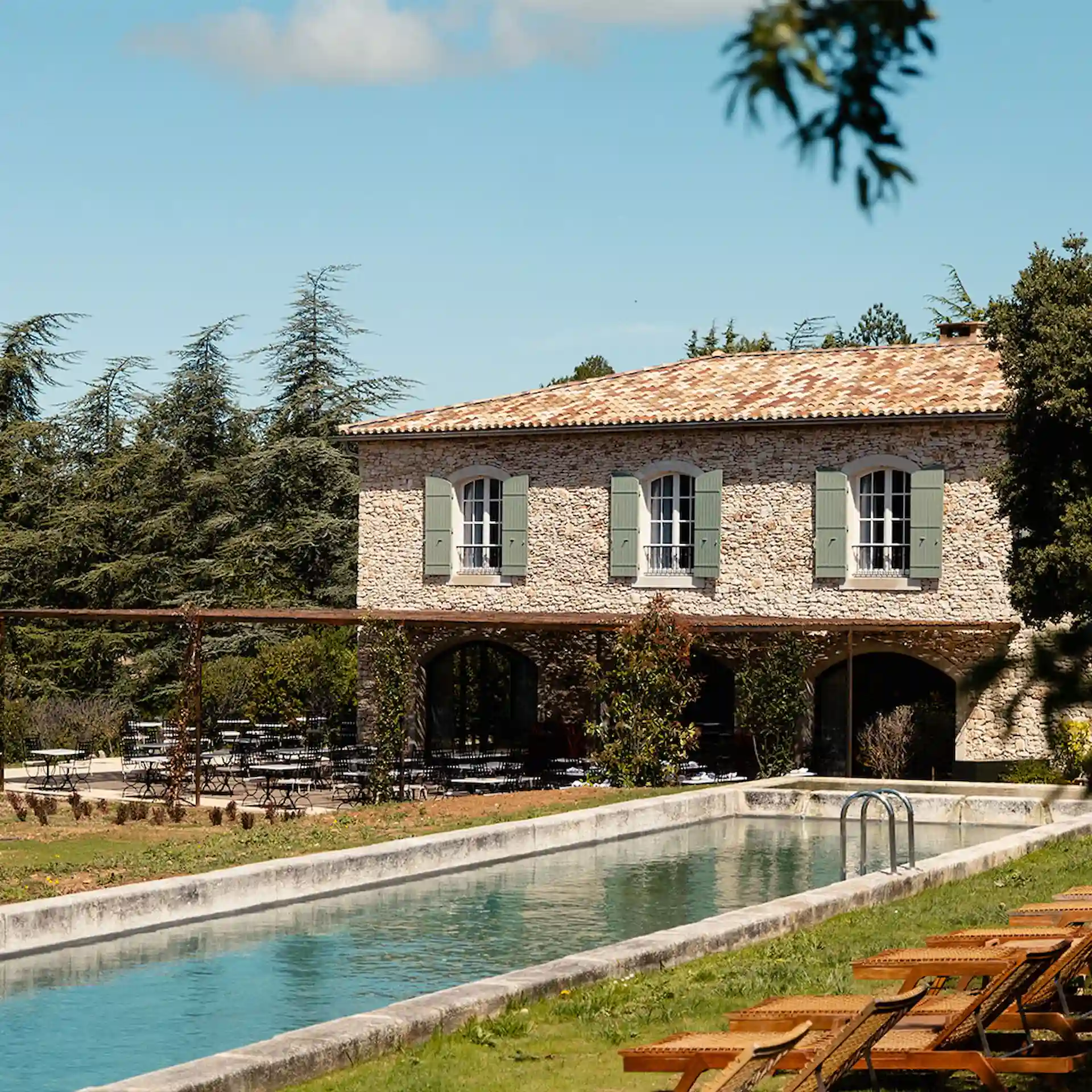 Stone countryside villa with green shutters, outdoor pool, lounge chairs, and garden seating under a clear blue sky.