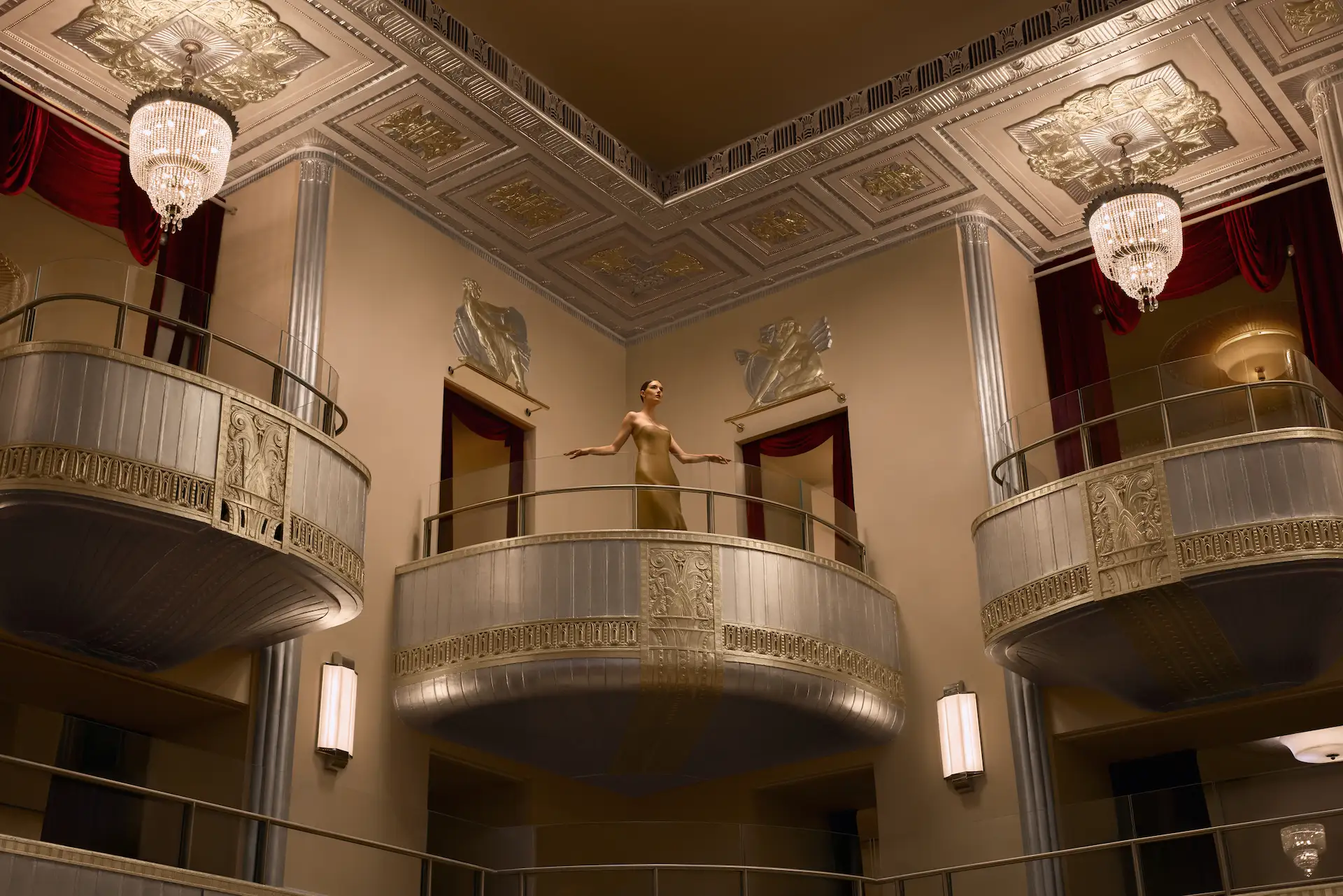 Woman in gold dress standing on ornate Art Deco balcony inside luxurious theater interior.
