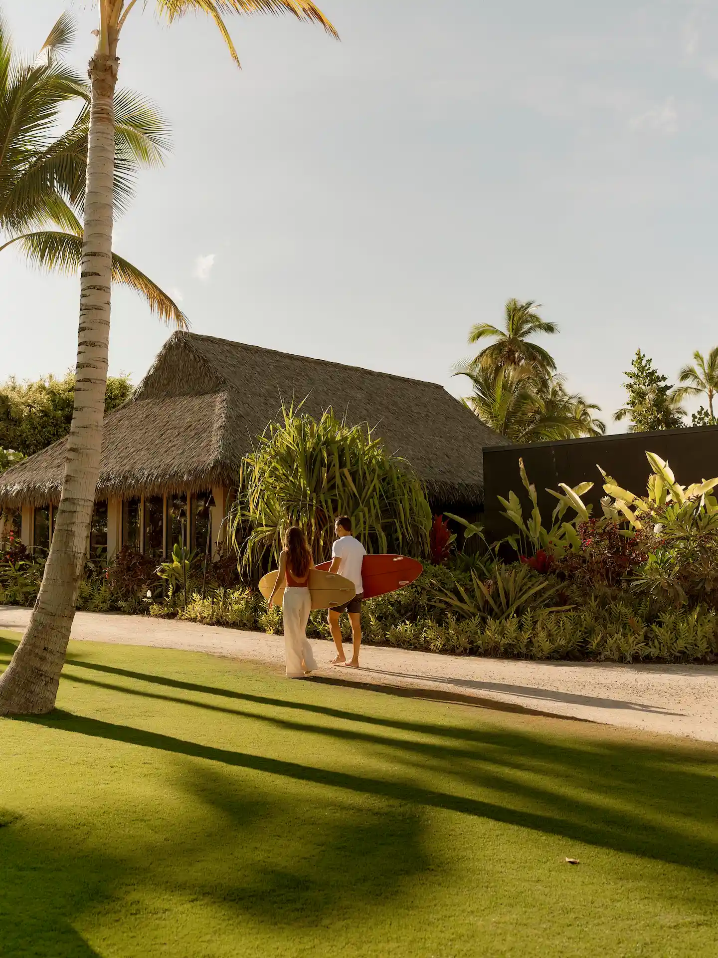 Tropical morning at Rosewood Kona Village with barefoot guests carrying surfboards past palm trees and thatched roof.