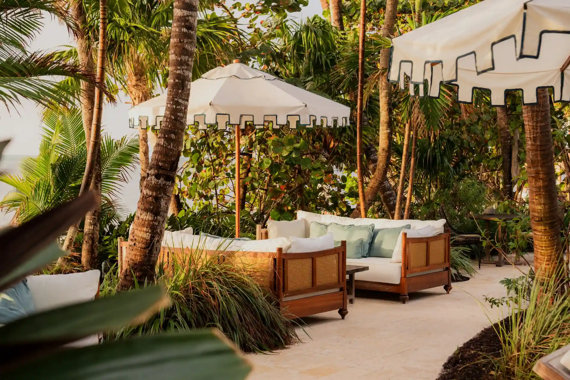 Outdoor lounge with white cushioned sofas, umbrellas, palm trees, and tropical landscaping.