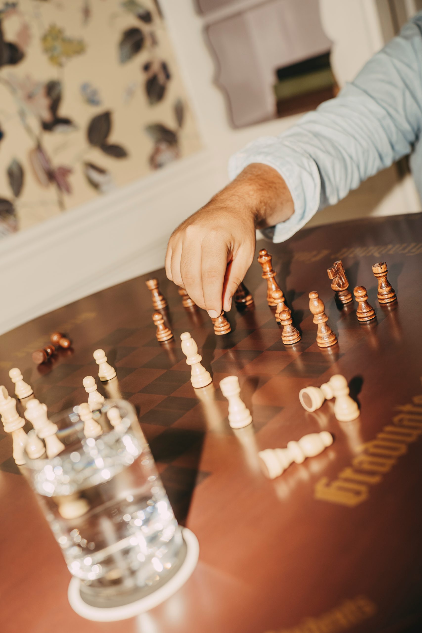 Person making a move in a chess game on a wooden board, with scattered pieces and a glass of water nearby.