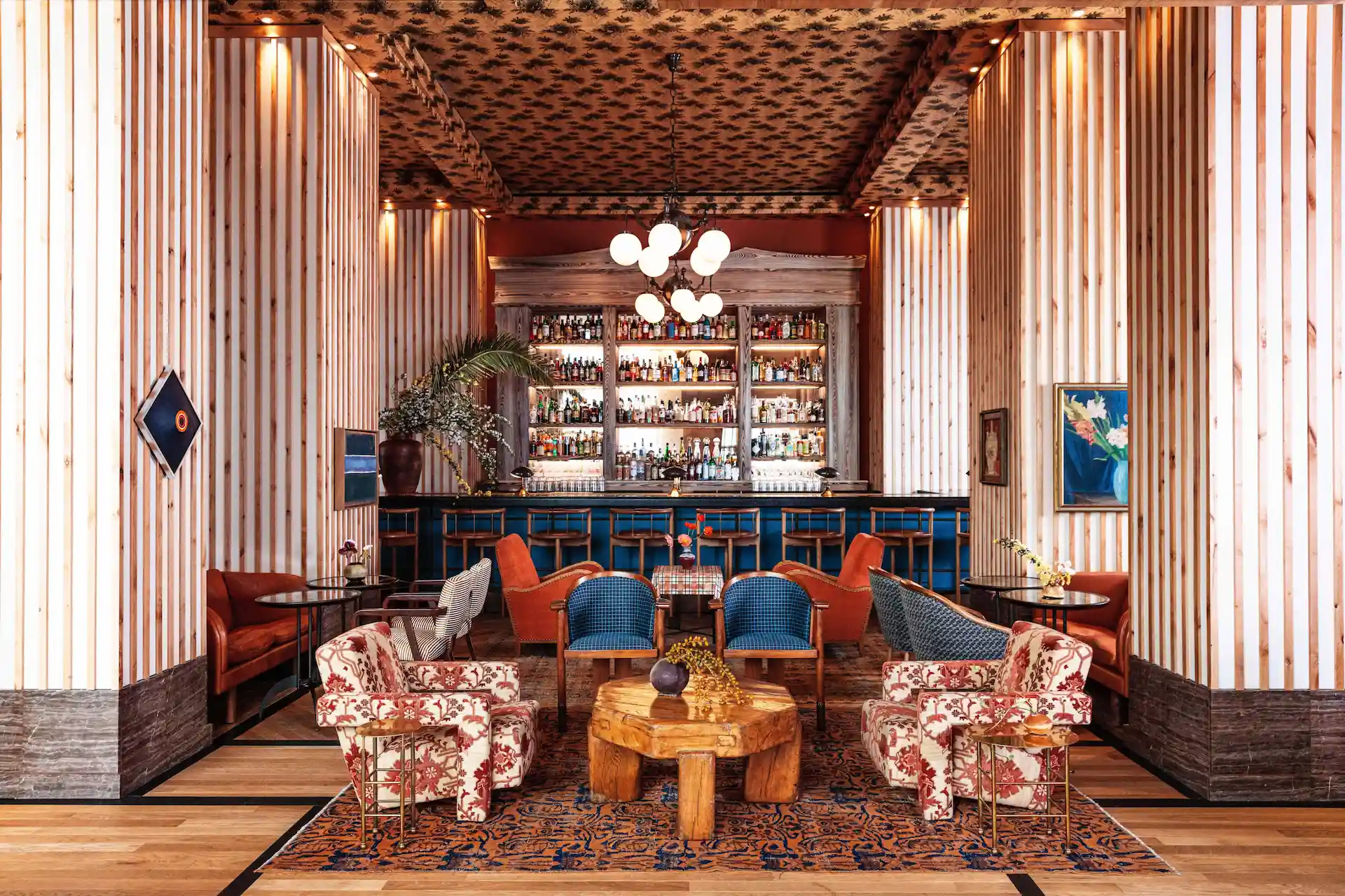 Proper Hotels 1 Eclectic bar interior with blue counter, vintage armchairs, patterned rug, and shelves of liquor bottles.