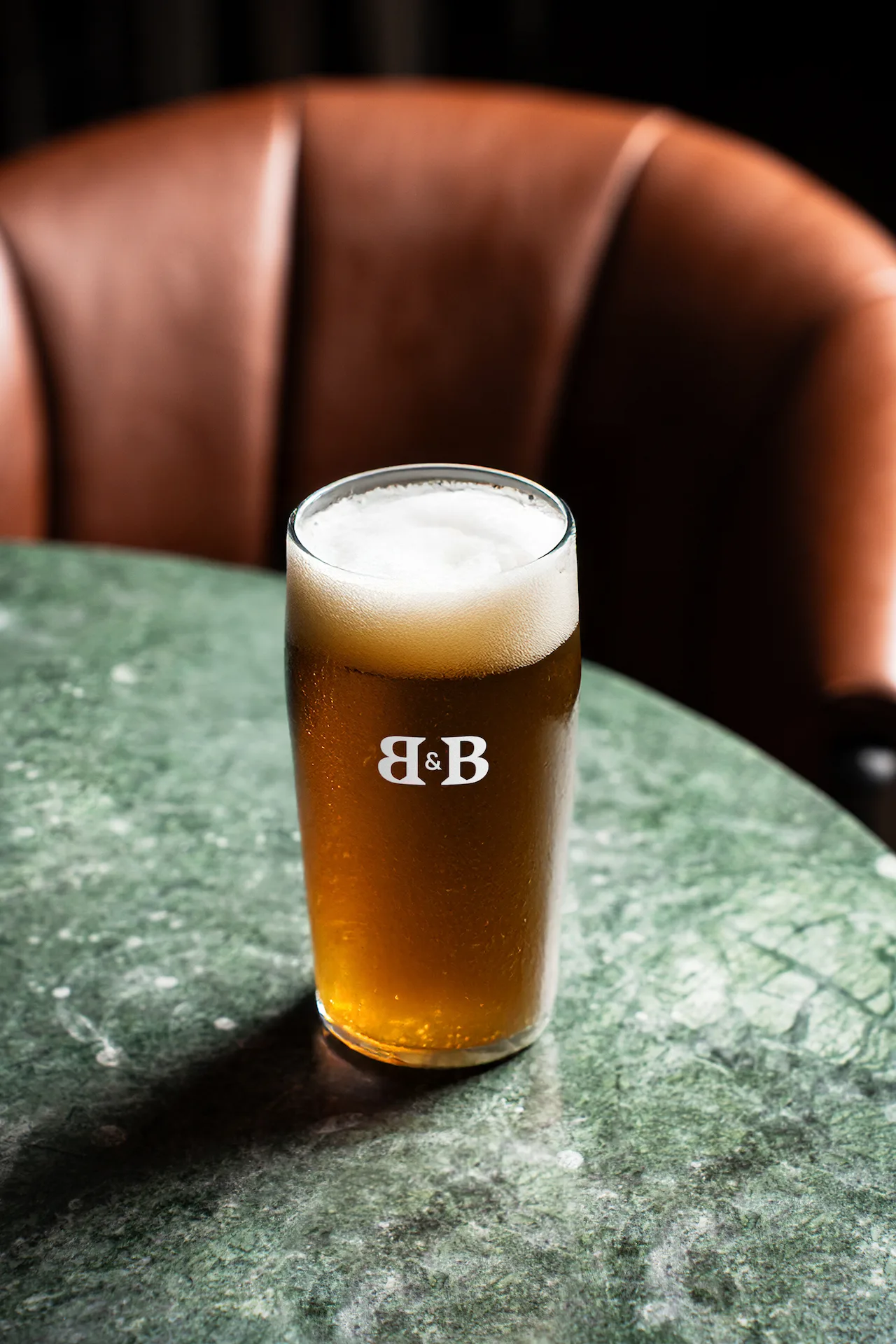 Pint glass of beer with “B & B” logo on green marble table, brown leather chair in background.