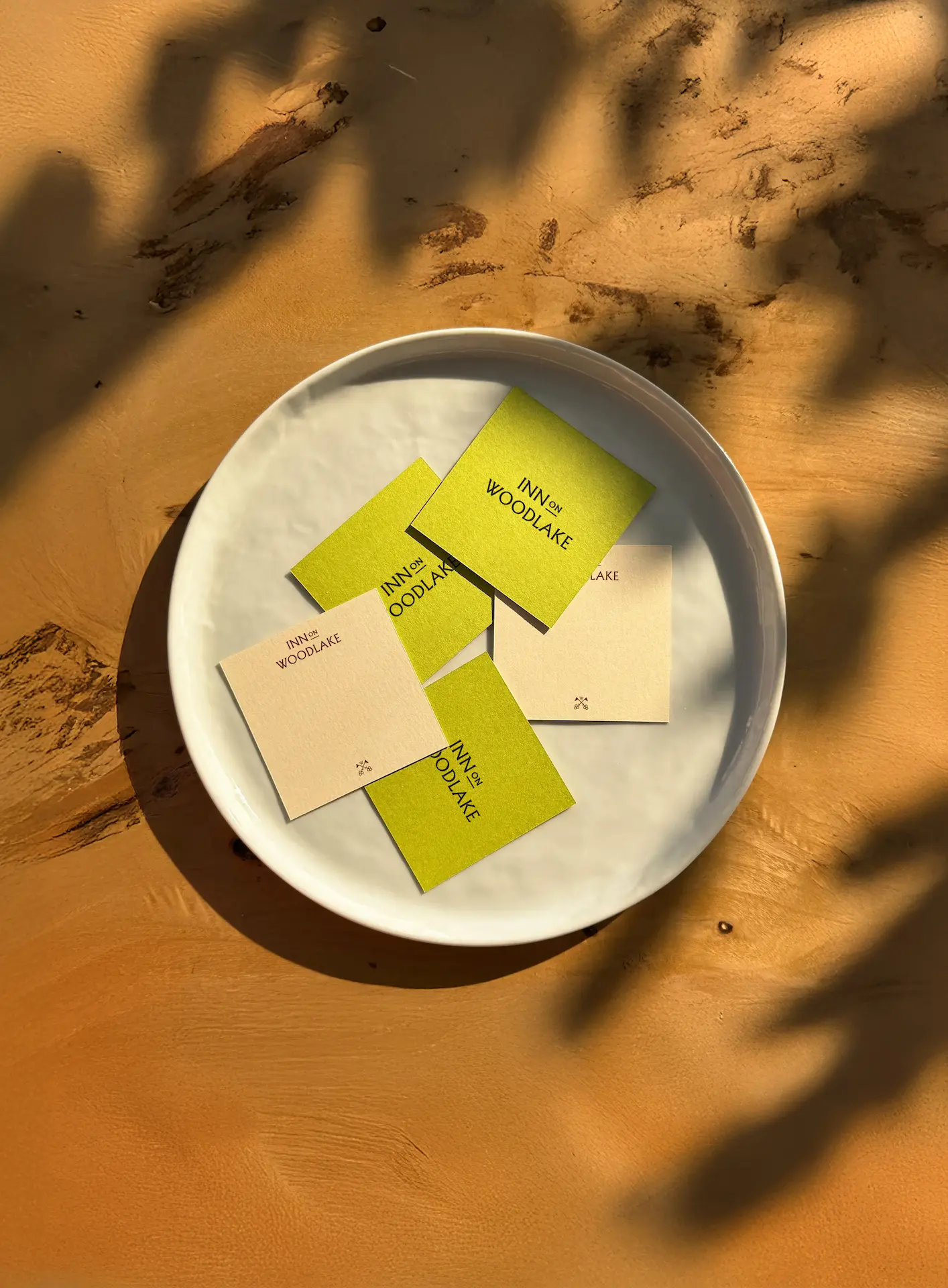 White ceramic dish on wooden surface with leaf shadows, holding five branded Inn on Woodlake cards in green and beige.