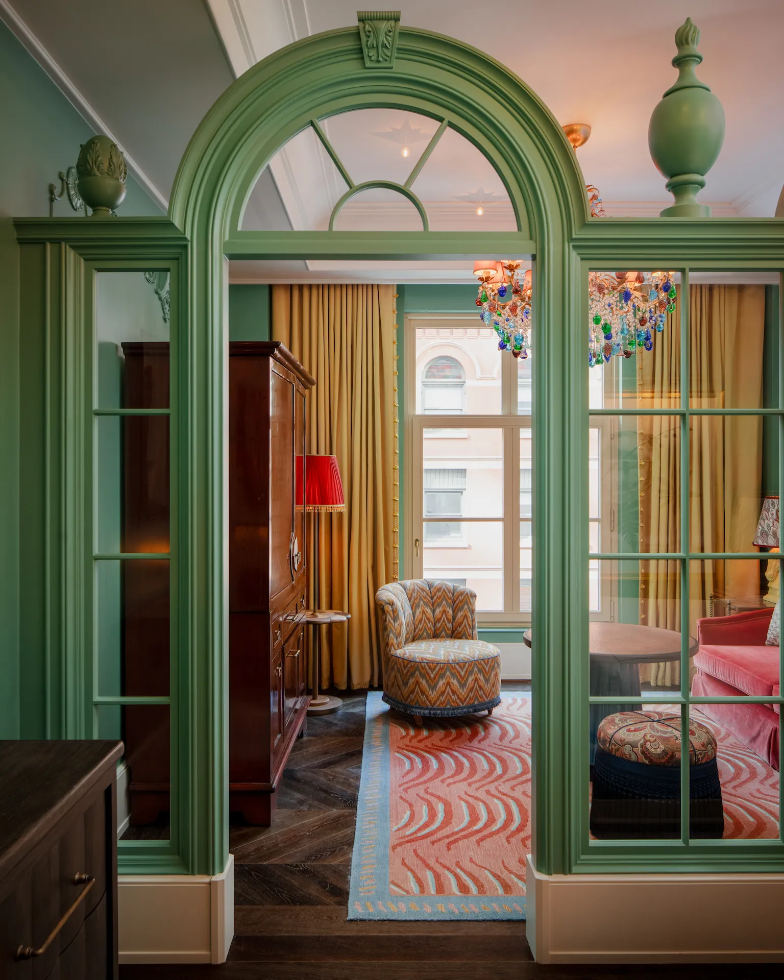 Colorful lounge with green archway, red velvet sofa, patterned armchair, and multicolored glass chandelier.