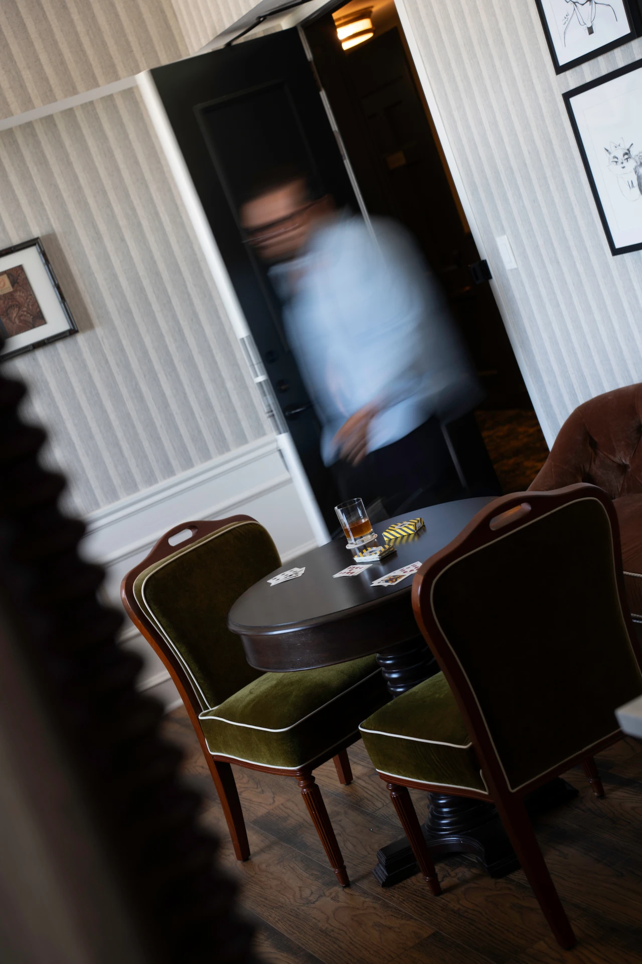 Round table with playing cards, drink, cigarettes, and green velvet chairs in vintage lounge setting.