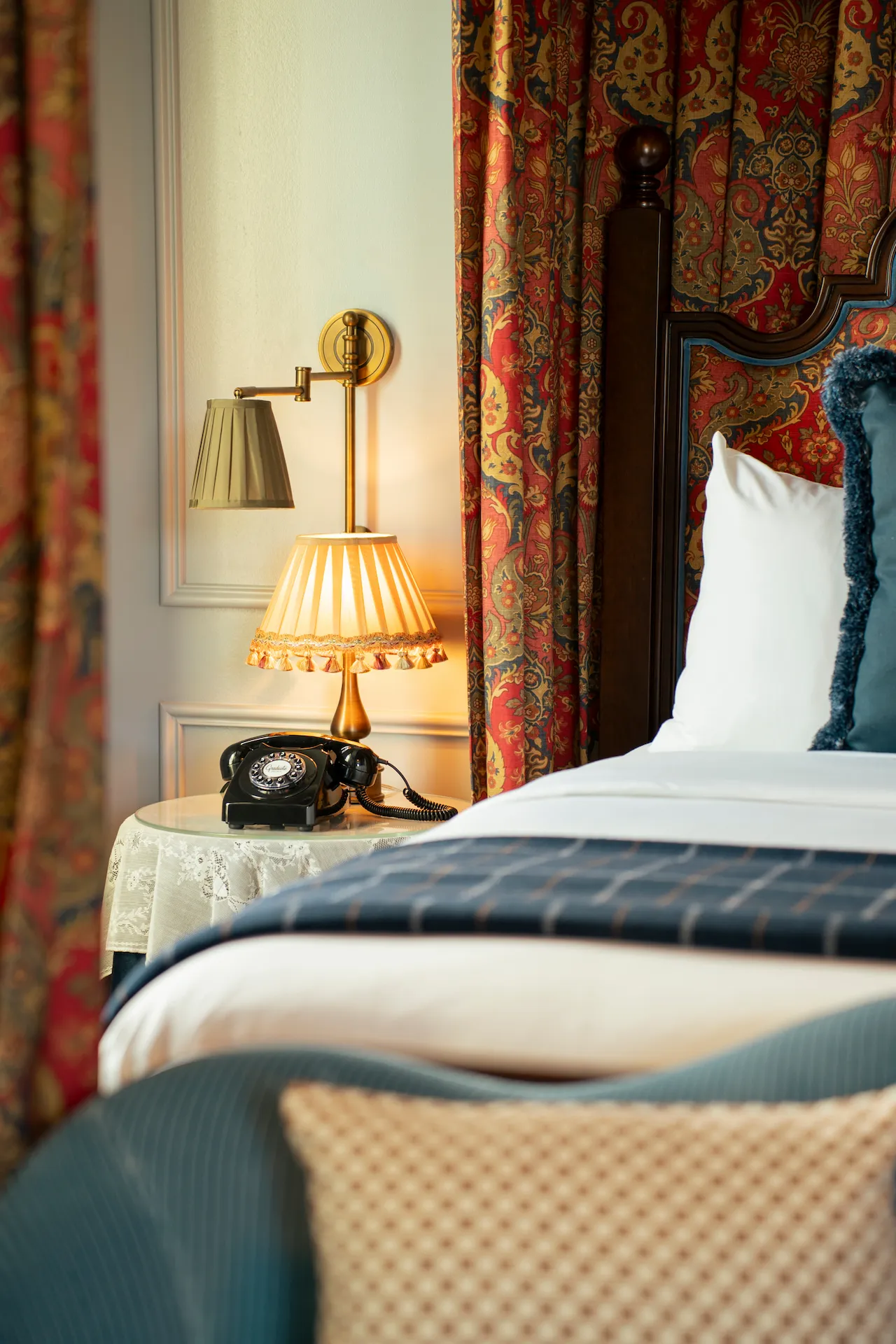 Vintage-style hotel bedroom corner with dark wood bed, lace-covered side table, rotary phone, and pleated lampshade.