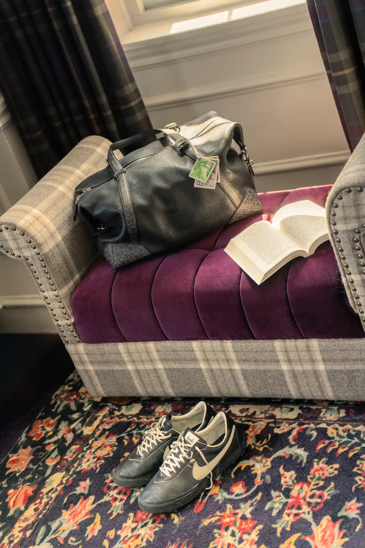 Plaid bench with velvet cushion, leather duffel bag, open book, and Nike sneakers on floral carpet.