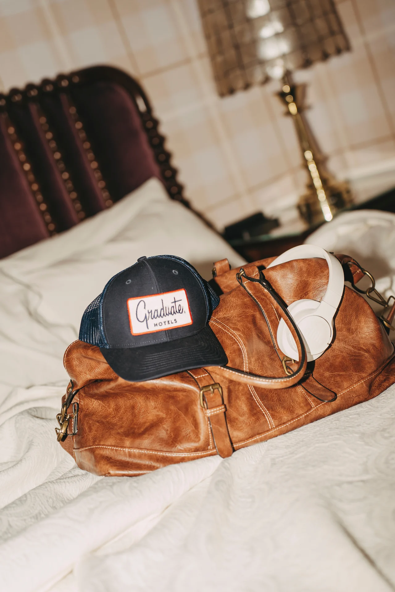 Brown leather duffel bag on hotel bed with Graduate Hotels cap and white headphones, purple headboard behind.
