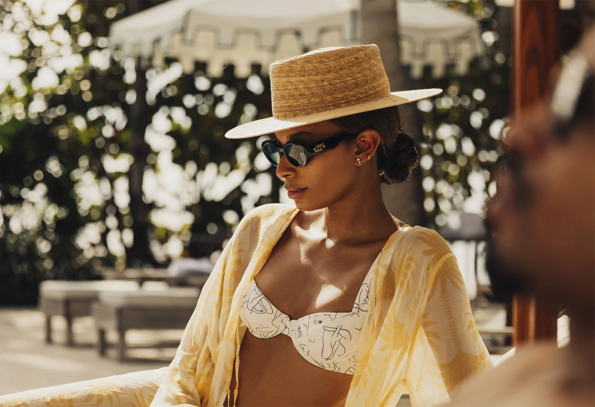 Woman in straw hat and abstract bikini lounging outdoors with sunlit umbrella and tropical backdrop.