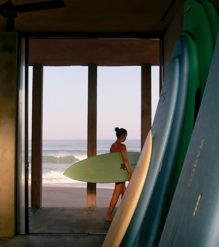 Person in red swimsuit holds green surfboard at entrance, looking toward ocean with stored boards nearby.