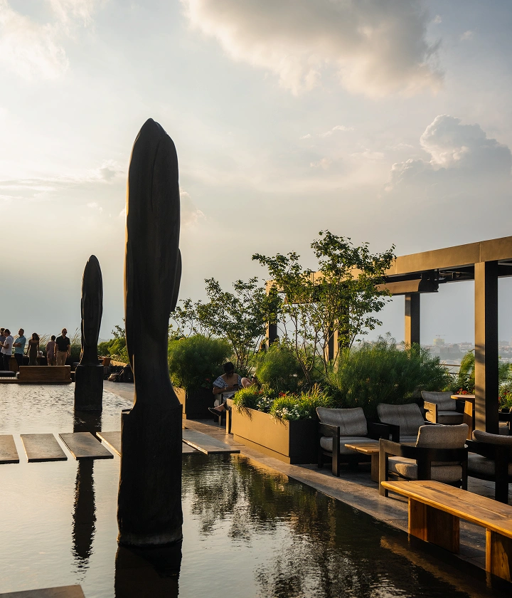 Rooftop lounge with water feature, sculptures, sunset light, and people gathered near terrace edge.