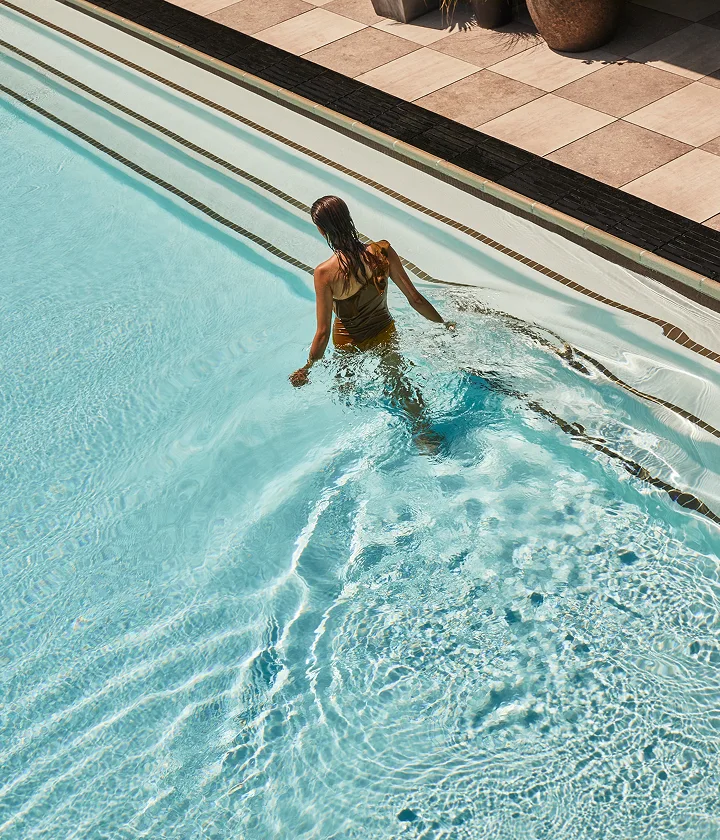 Person in swimsuit steps into clear blue pool with tiled edges and sunlit ripples.
