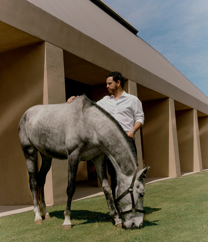 Man in white shirt stands beside grazing horse near modern building with columns and slanted roof.