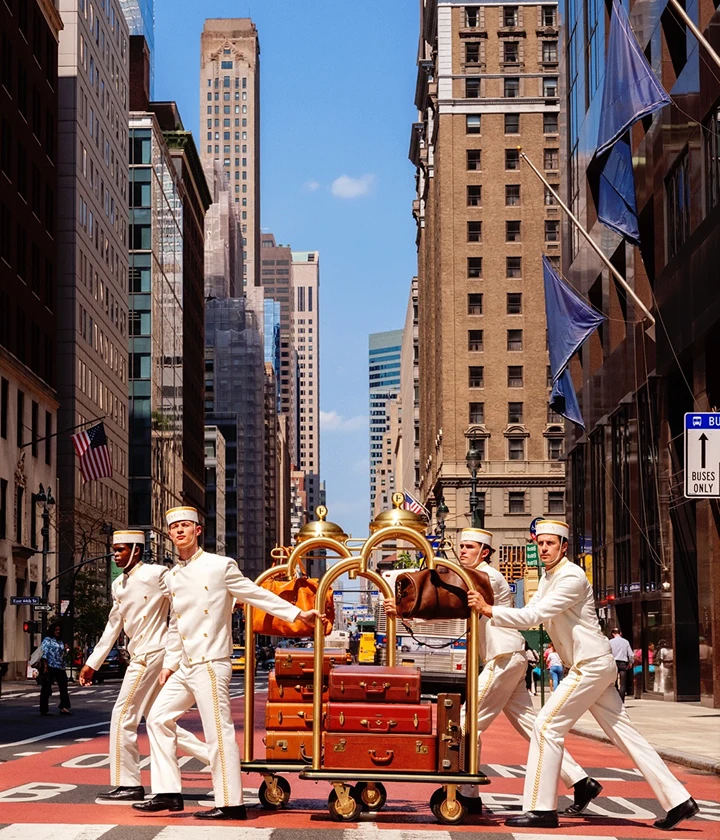 Bellhops in white uniforms push luggage carts across city street in choreographed formation.