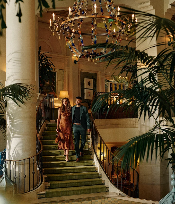 Couple descends ornate staircase under chandelier in elegant interior with columns, plants, and artwork.