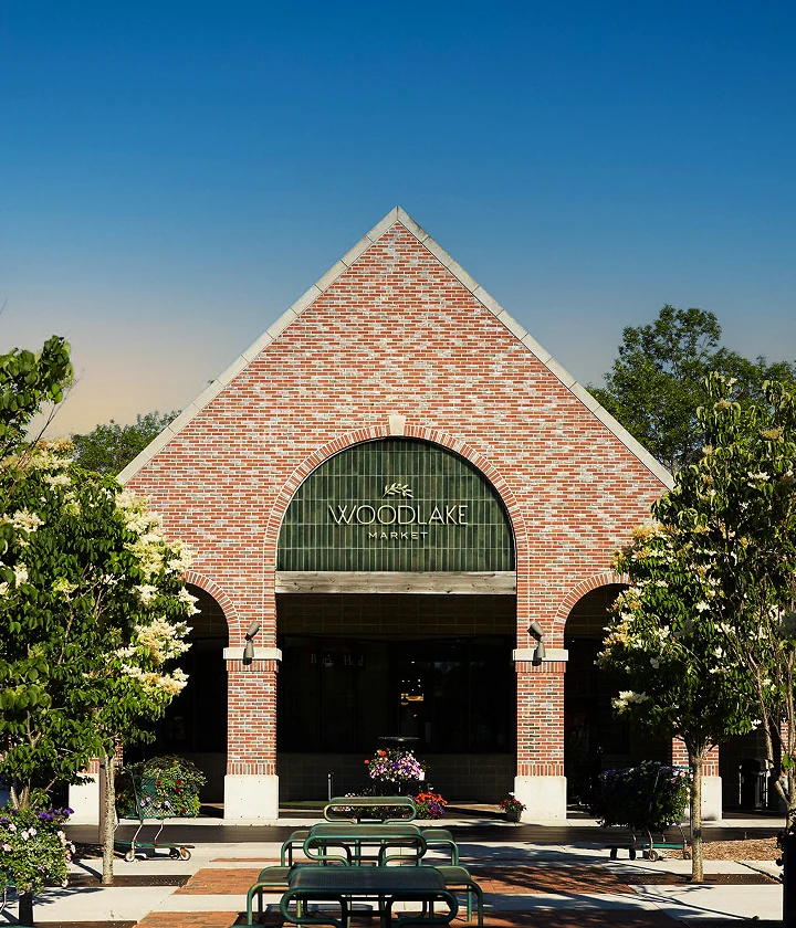 Branding by Spherical 17 Brick building with arched window labeled Woodlake Market, outdoor seating, flower pots, and trees.