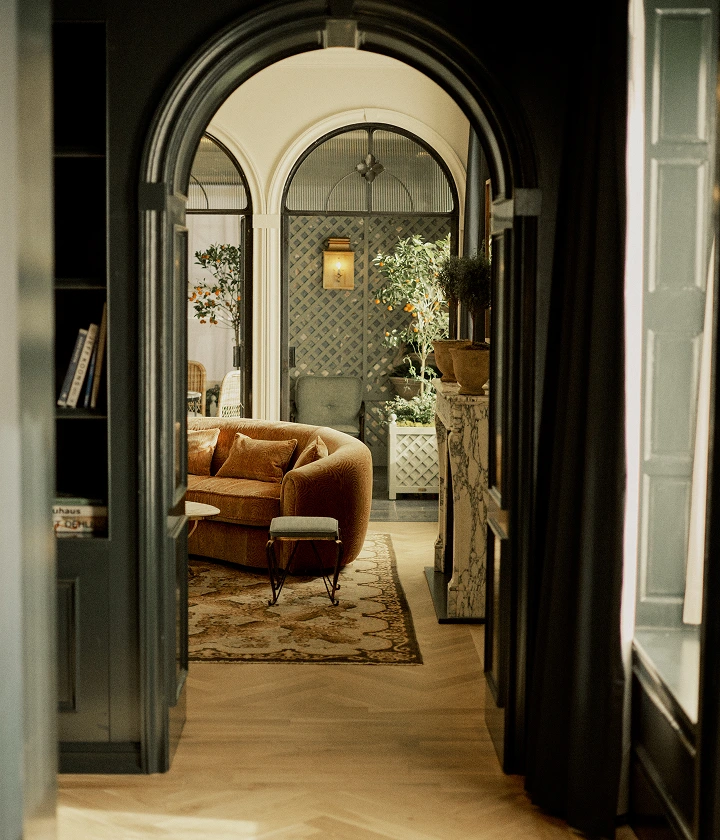 Velvet sofa and potted orange trees seen through arched doorway with bookshelves and wood floor.