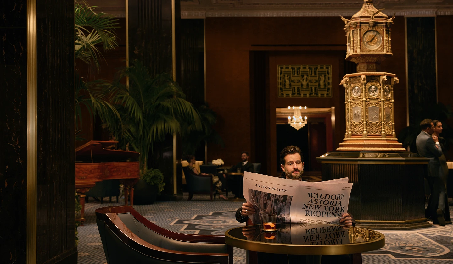 Man reading newspaper in elegant Waldorf Astoria lobby with grand clock, chandelier, and piano.