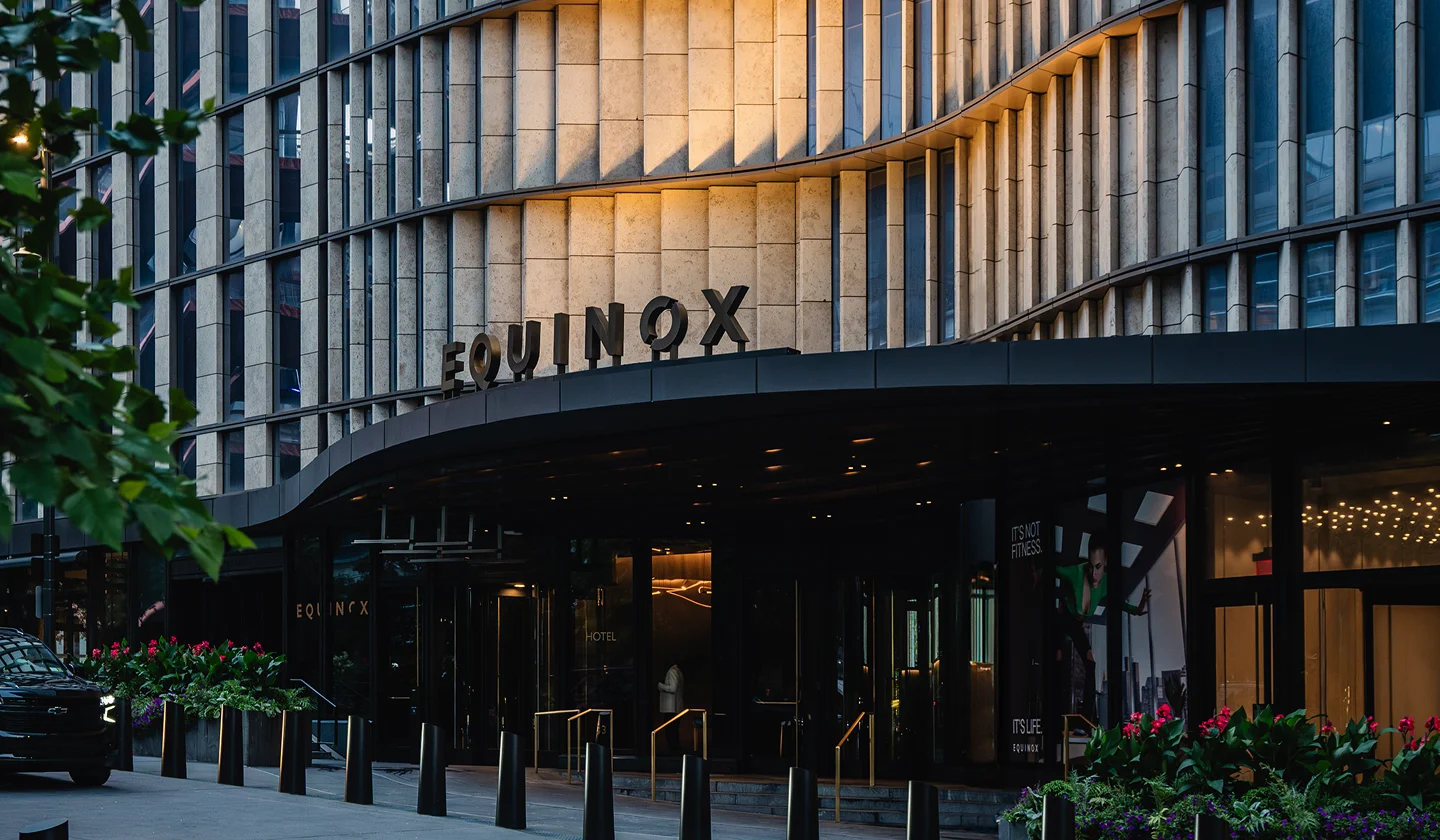 Equinox building with curved stone-and-glass facade, bold signage, and landscaped entrance with flowers.