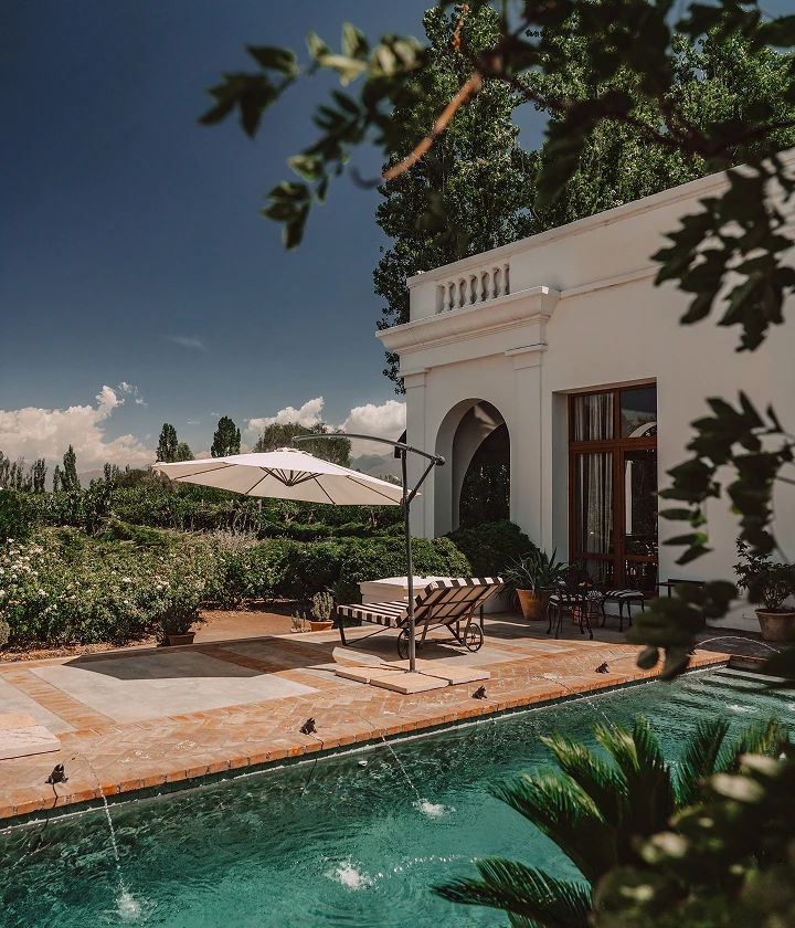 Poolside lounge with striped chaise, umbrella, water jets, and white building framed by greenery.