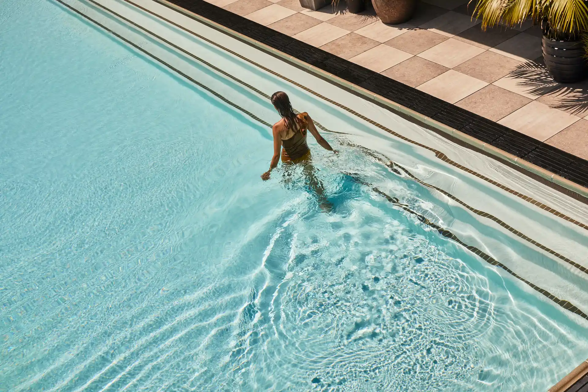 Proper Hotels 4 Person entering a clear blue pool, creating ripples, with tiled edges and potted plants in serene setting.