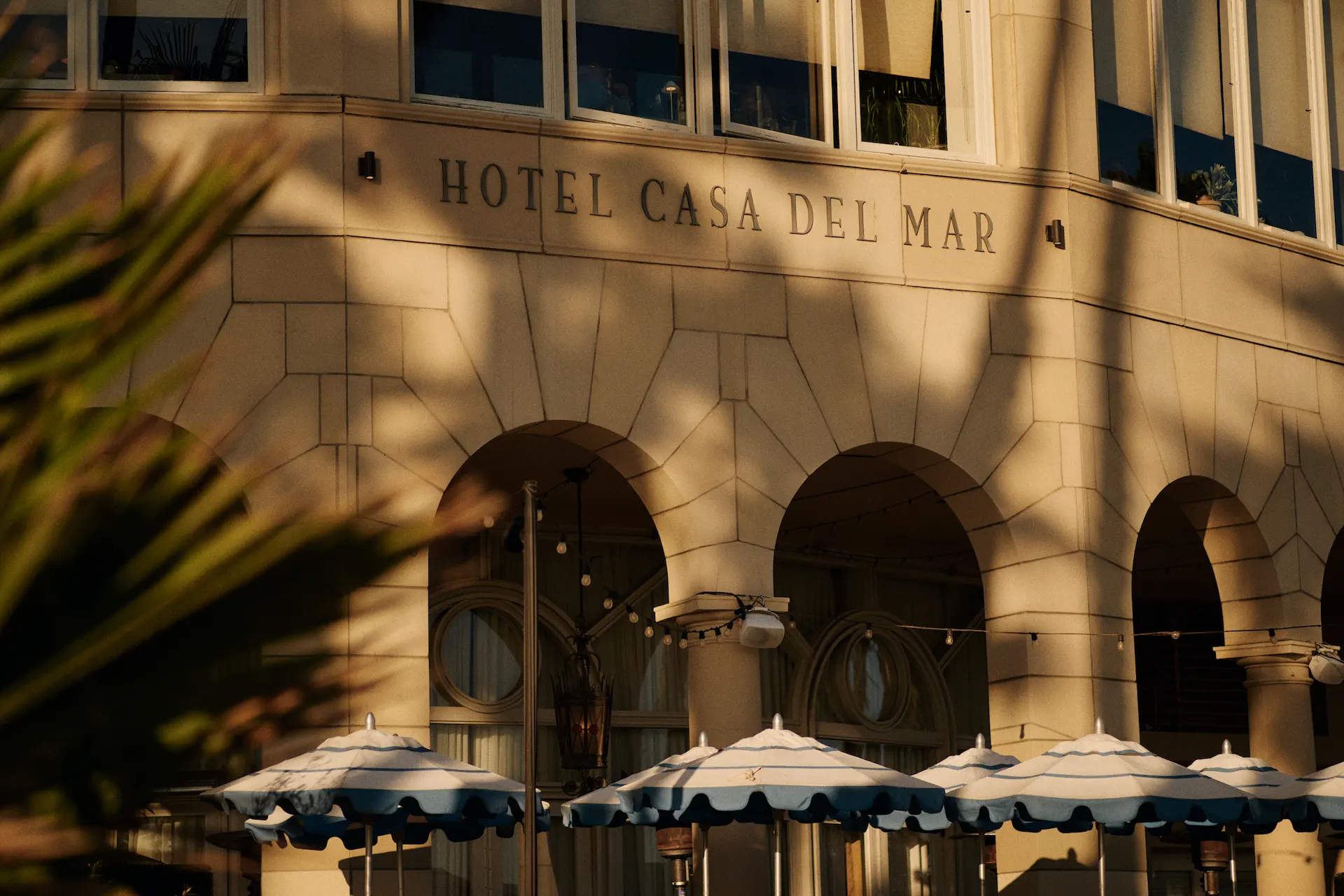 Facade of Hotel Casa Del Mar with arched windows, striped umbrellas, and palm fronds in warm coastal light.
