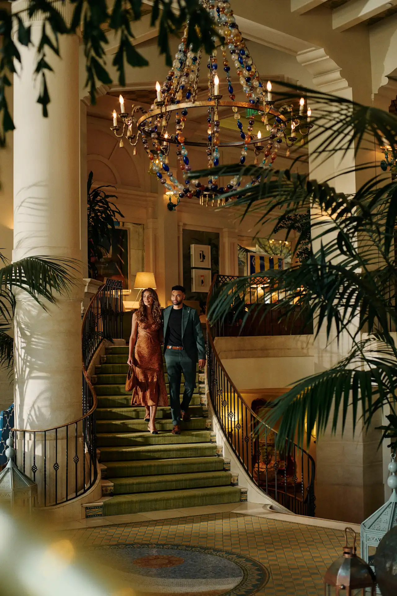Couple descending grand staircase at Casa Del Mar in Santa Monica Beach, beneath chandelier and surrounded by elegant decor.