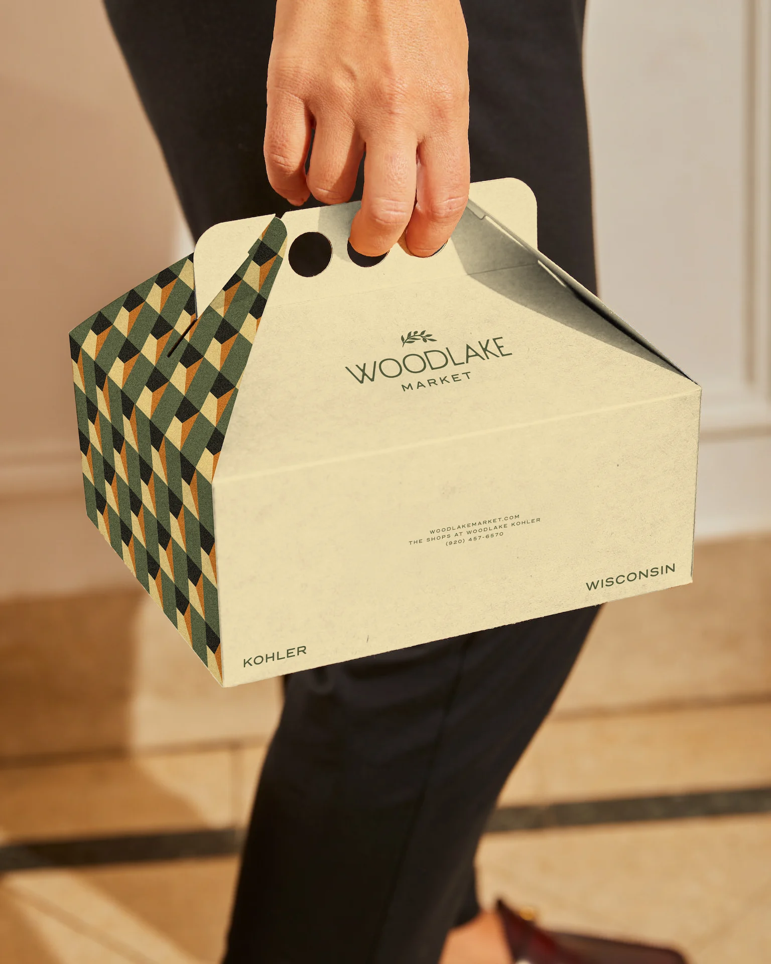 Woodlake Market 3 Person holding beige takeaway box with geometric pattern and Woodlake Market branding, including website and contact info.