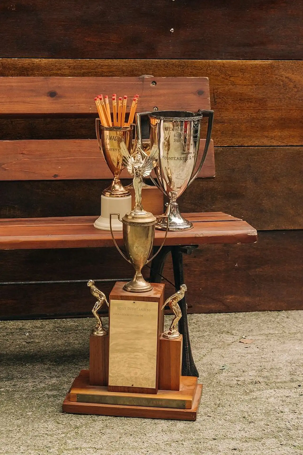 Four varied trophies on wooden bench, one filled with pencils; engraved with award titles and contest details.